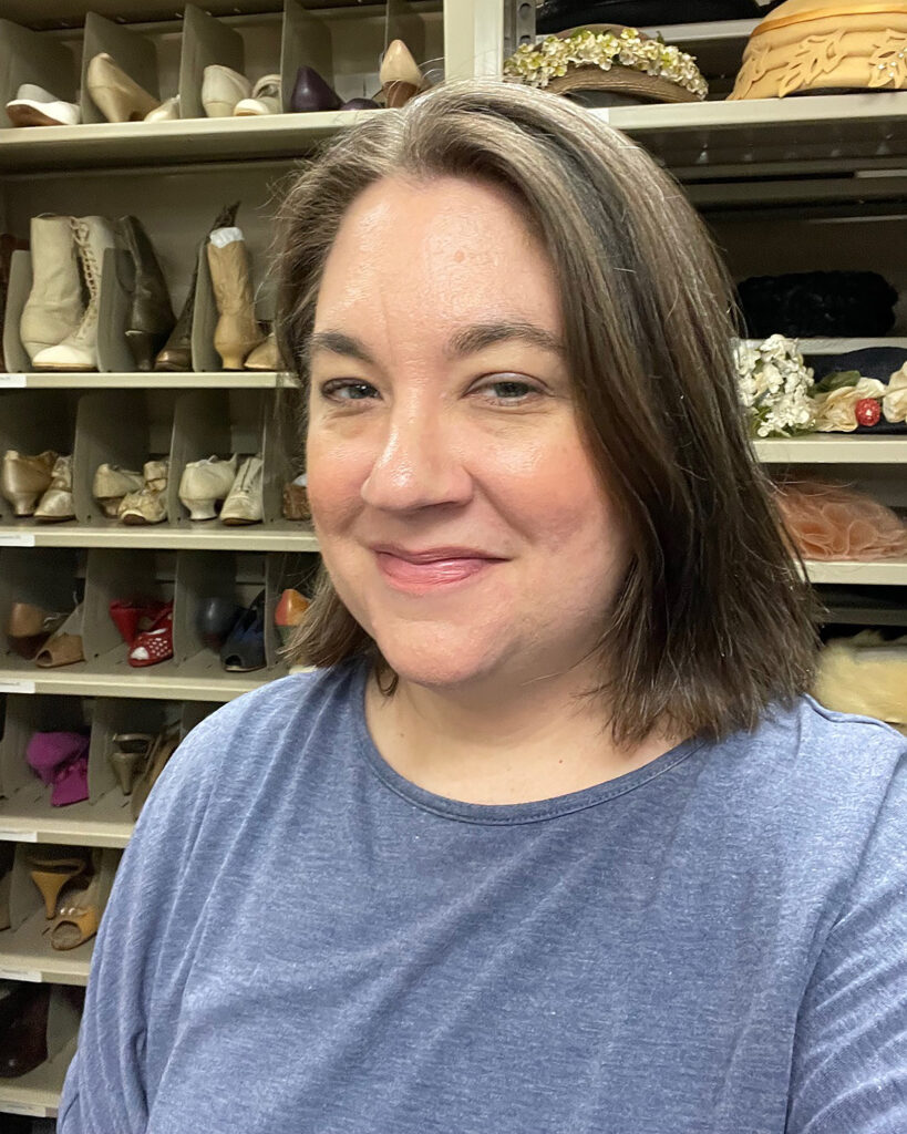 Brown, haired female wearing a blue shirt standing in front of shelves with hats and shoes on them.
